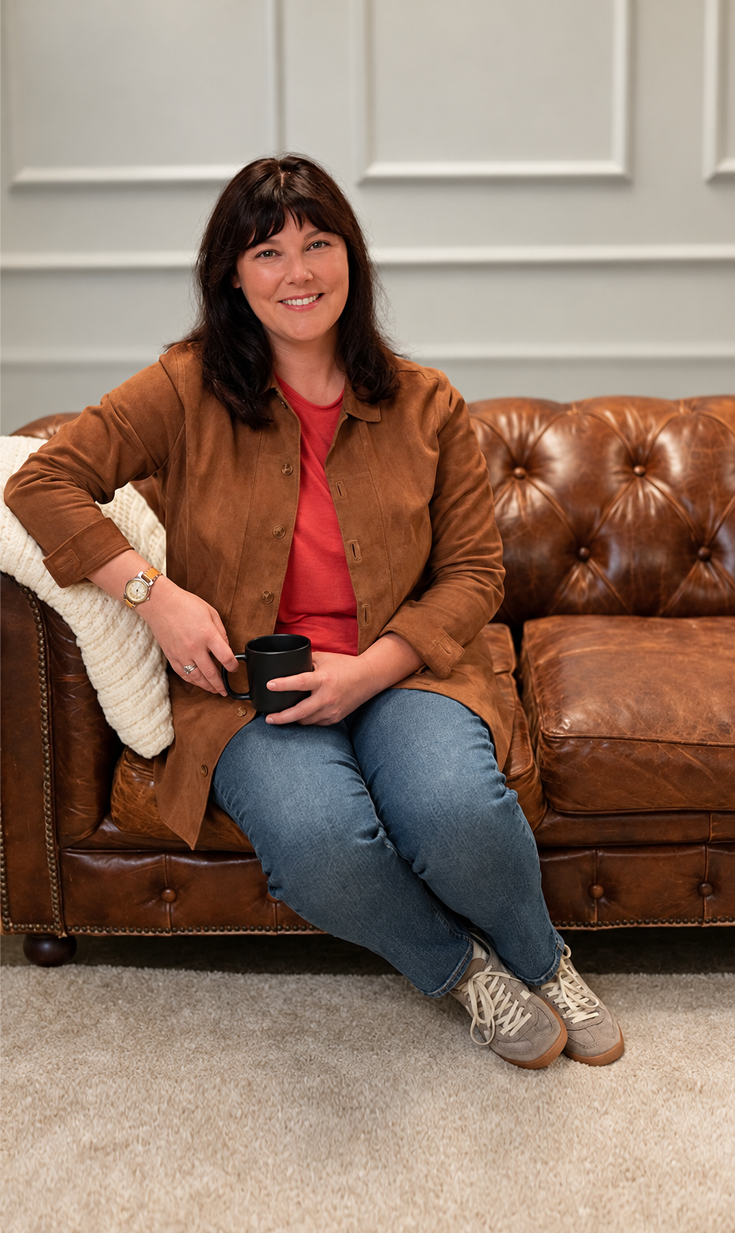 Jess Lorimer, founder of Selling to Corporate, seated on a leather sofa holding a mug.